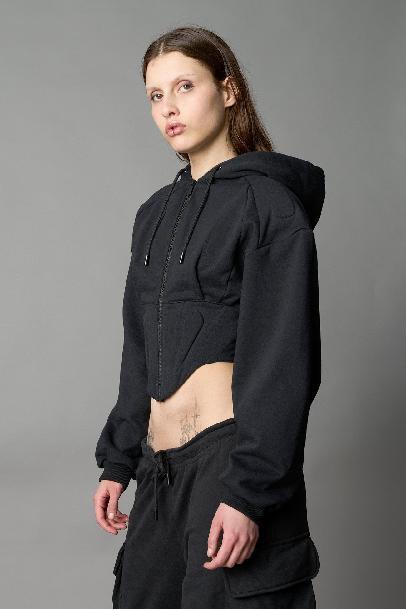 model in a black cropped hoodie featuring structured corset-style paneling embroidered across the chest in tonal black