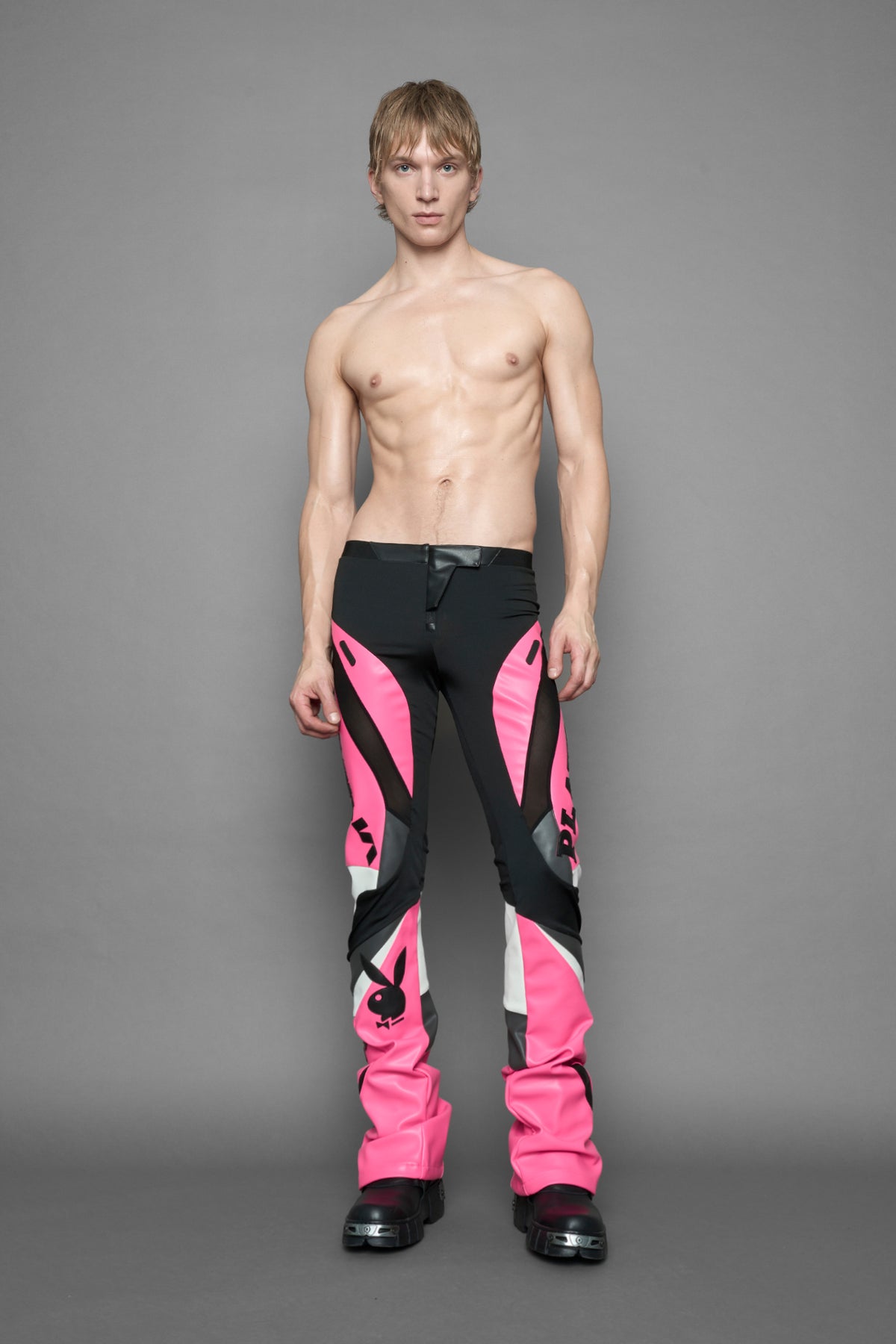 A female model wearing a pink and black form fitted Pants with PU Leather Embroidery of 'Namilia', 'Playboy'. The pants have black Mesh fabric and dark grey PU leather panels and are slightly flared from knee to hem.