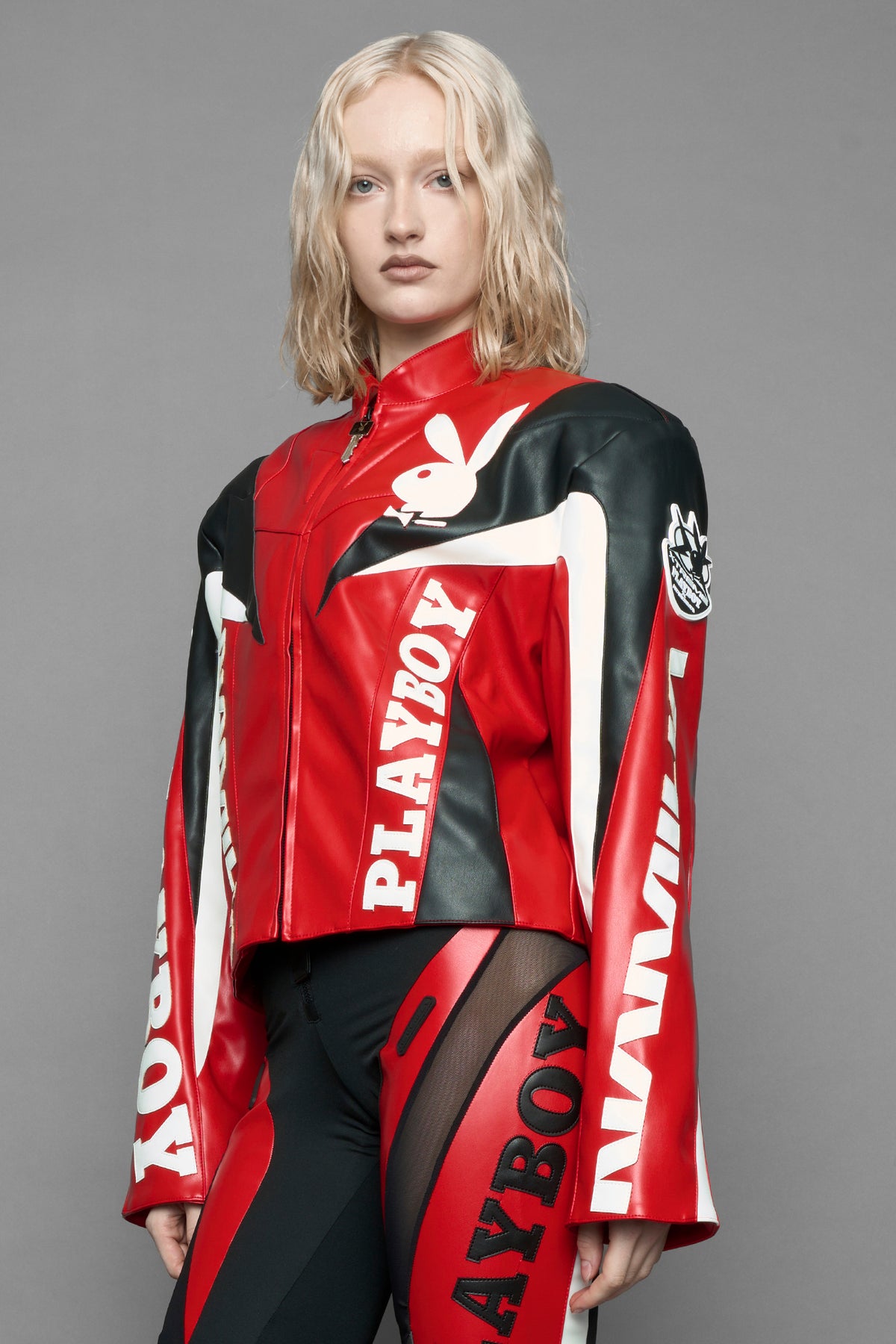 model wearing red vegan leather zip-up jacket with black PU leather 'Namilia' and 'Playboy' embroidery and black and white paneling. On the left chest there is a white Playboy Bunny Embroidery.