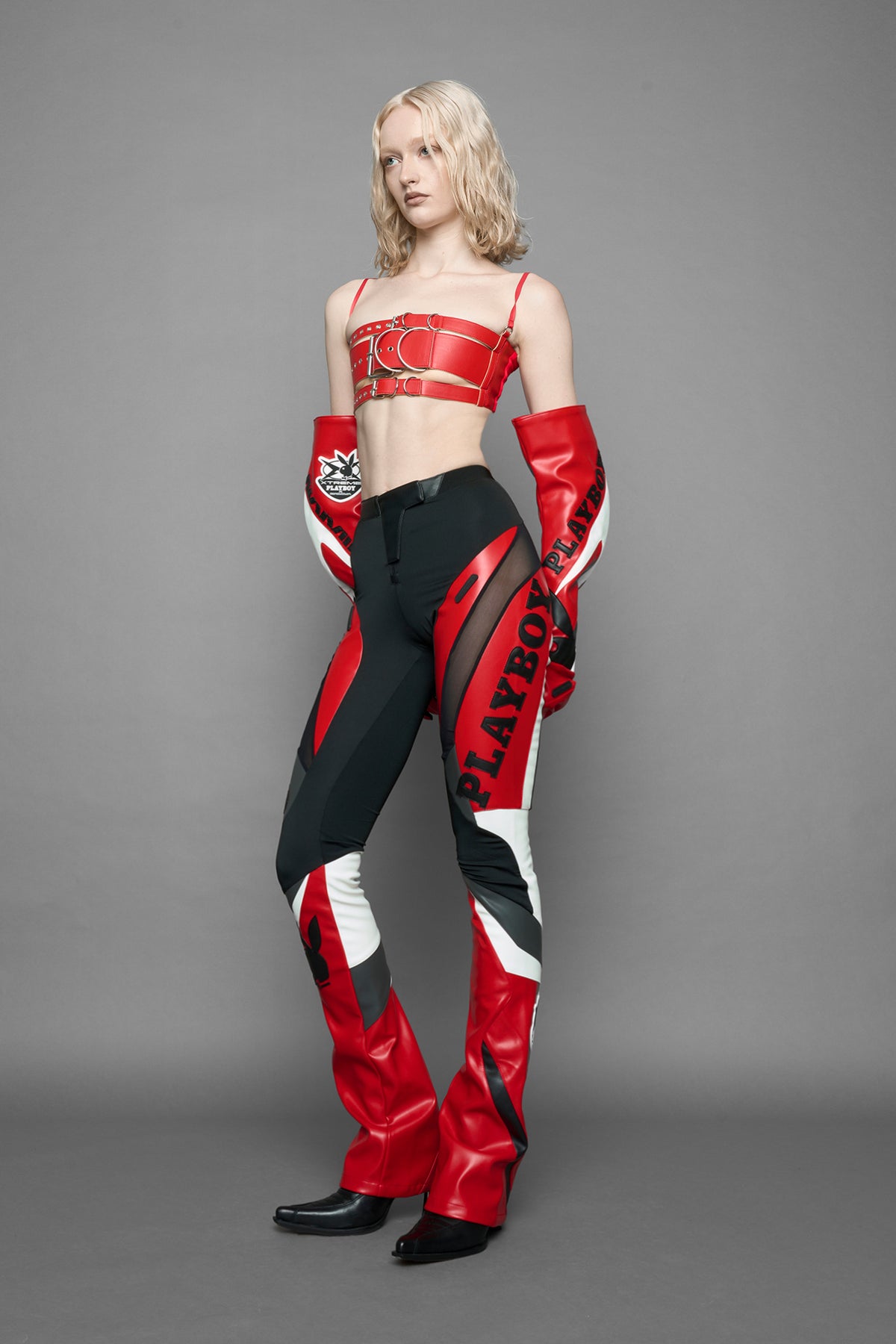 model wearing a red and black form fitted Pants with PU Leather Embroidery of 'Namilia', 'Playboy'. The pants have black Mesh fabric and dark grey PU leather panels and are slightly flared from knee to hem