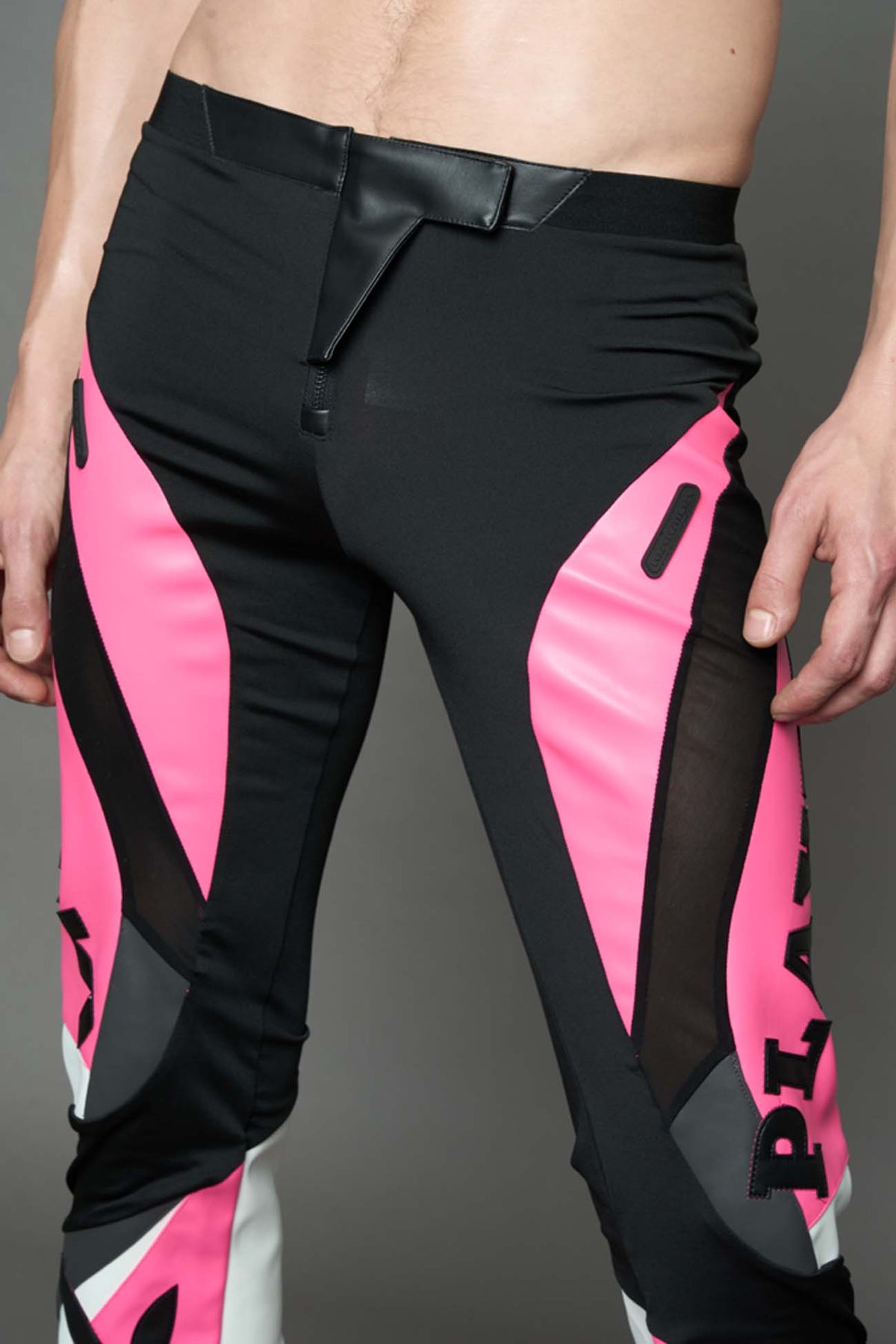 A female model wearing a pink and black form fitted Pants with PU Leather Embroidery of 'Namilia', 'Playboy'. The pants have black Mesh fabric and dark grey PU leather panels and are slightly flared from knee to hem.