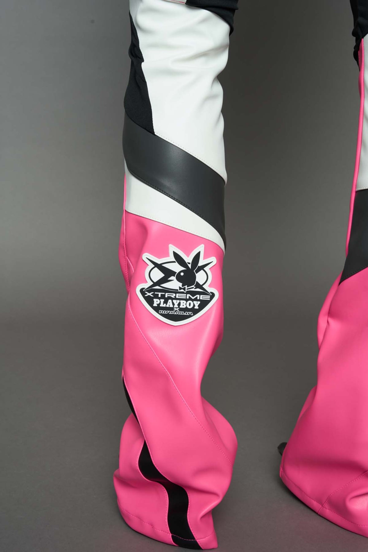A female model wearing a pink and black form fitted Pants with PU Leather Embroidery of 'Namilia', 'Playboy'. The pants have black Mesh fabric and dark grey PU leather panels and are slightly flared from knee to hem.