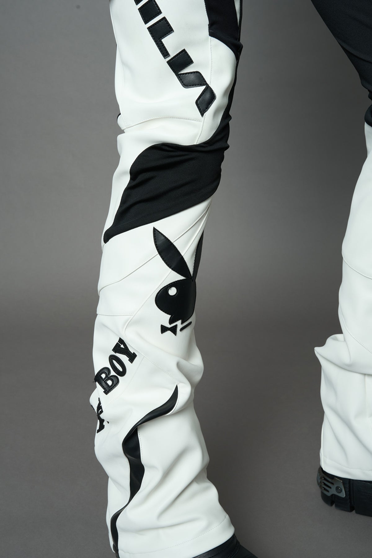A female model wearing a white and black form fitted Pants with PU Leather Embroidery of 'Namilia', 'Playboy'. The pants have black Mesh fabric panels and are slightly flared from knee to hem