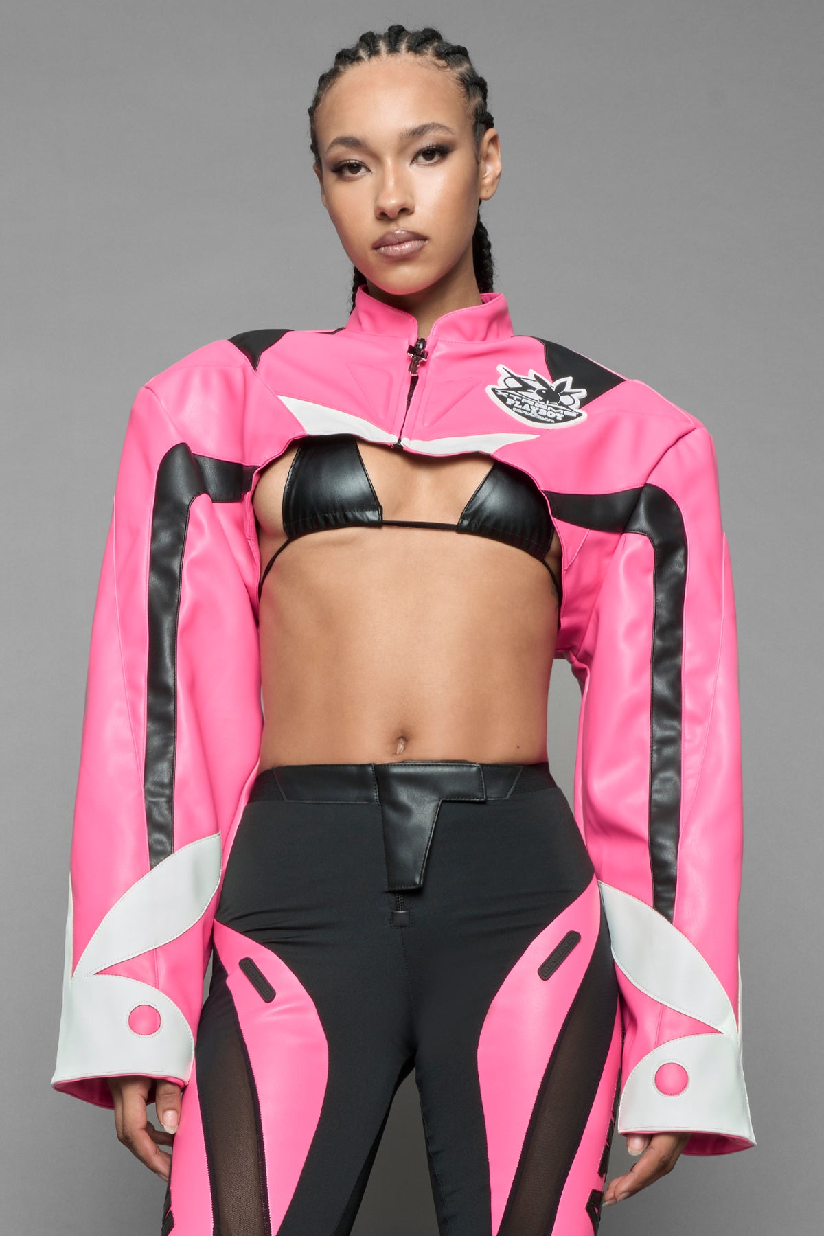 A female model wearing a vegan leather cropped jacket with broad shoulders in pink with black and white paneling. The jacket has PU leather embroidery of 'Namilia' and 'Playboy' with a key formed zipper pull and a rubbertag on the left breast.