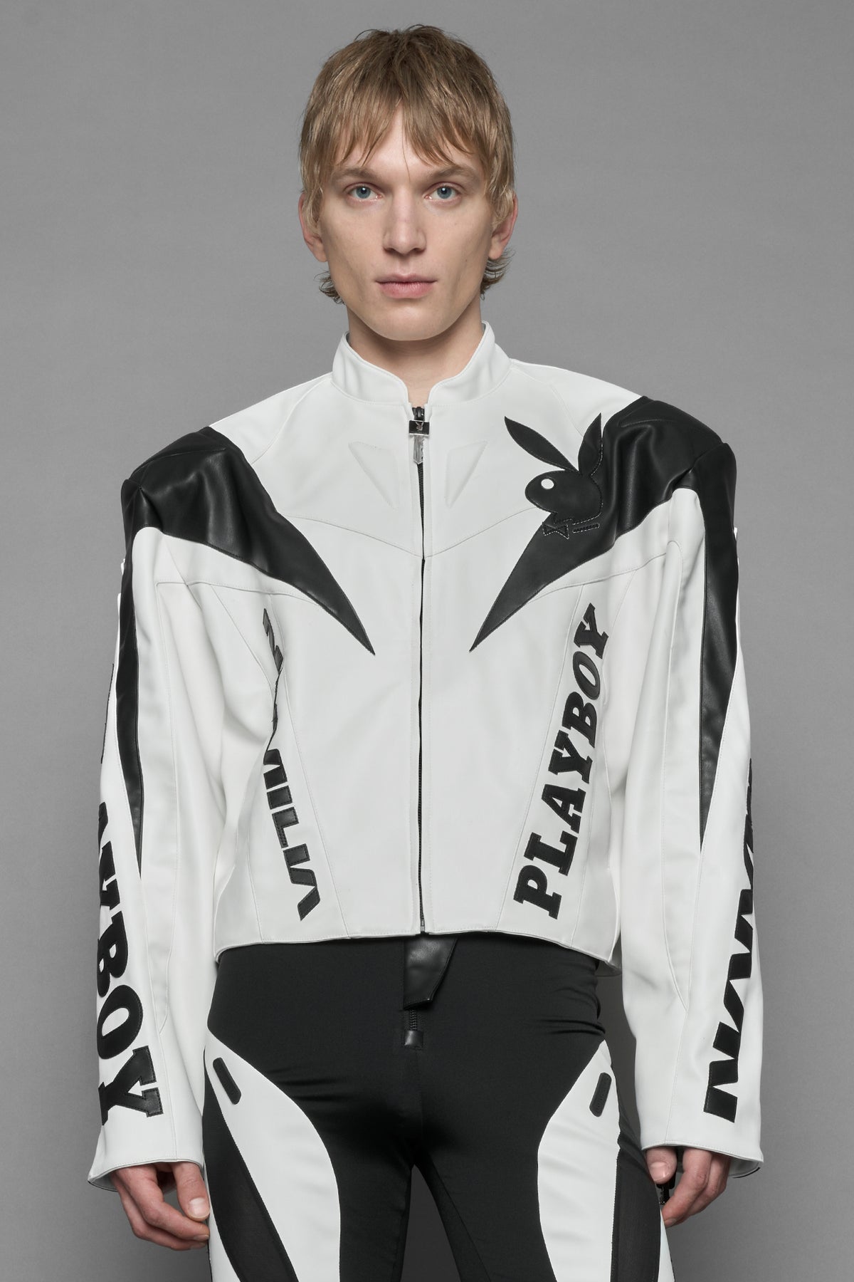 A male model wearing white vegan leather zip-up jacket with black PU leather 'Namilia' and 'Playboy' embroidery and black and white paneling. On the left chest there is a white Playboy Bunny Embroidery.