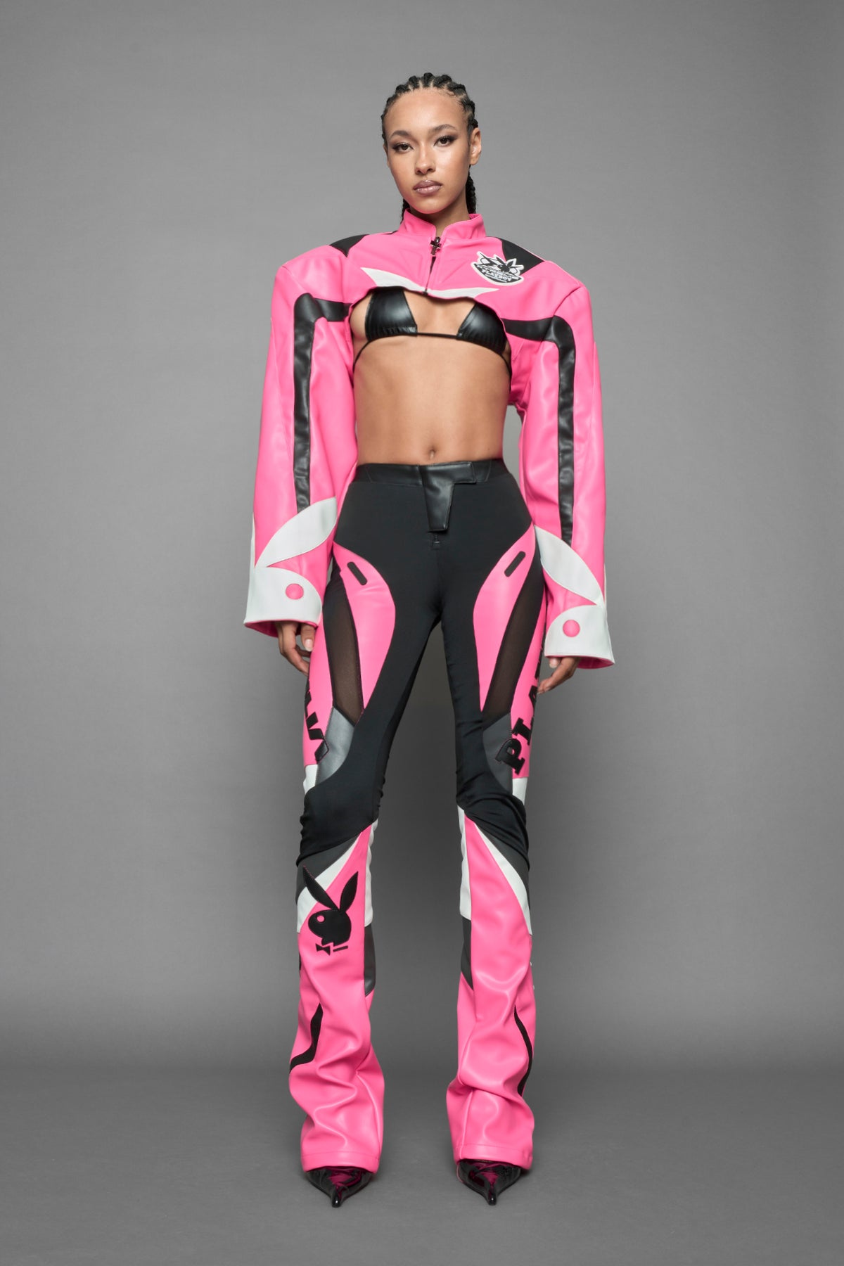 A female model wearing a pink and black form fitted Pants with PU Leather Embroidery of 'Namilia', 'Playboy'. The pants have black Mesh fabric and dark grey PU leather panels and are slightly flared from knee to hem.