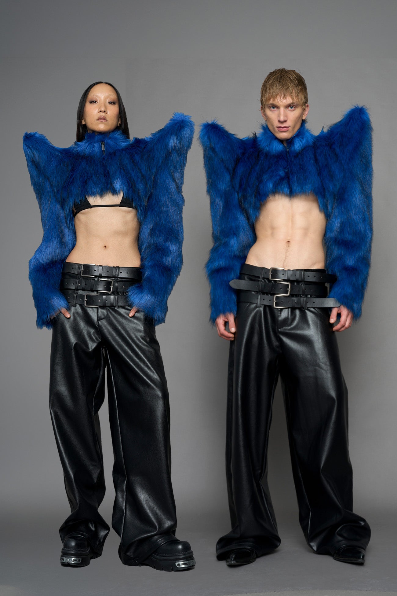 Two models standing side by side in a studio setting, wearing matching royal blue furry cropped jackets with dramatic wing-like shoulders, black leather-like wide-leg pants, and black belts against a plain gray background.