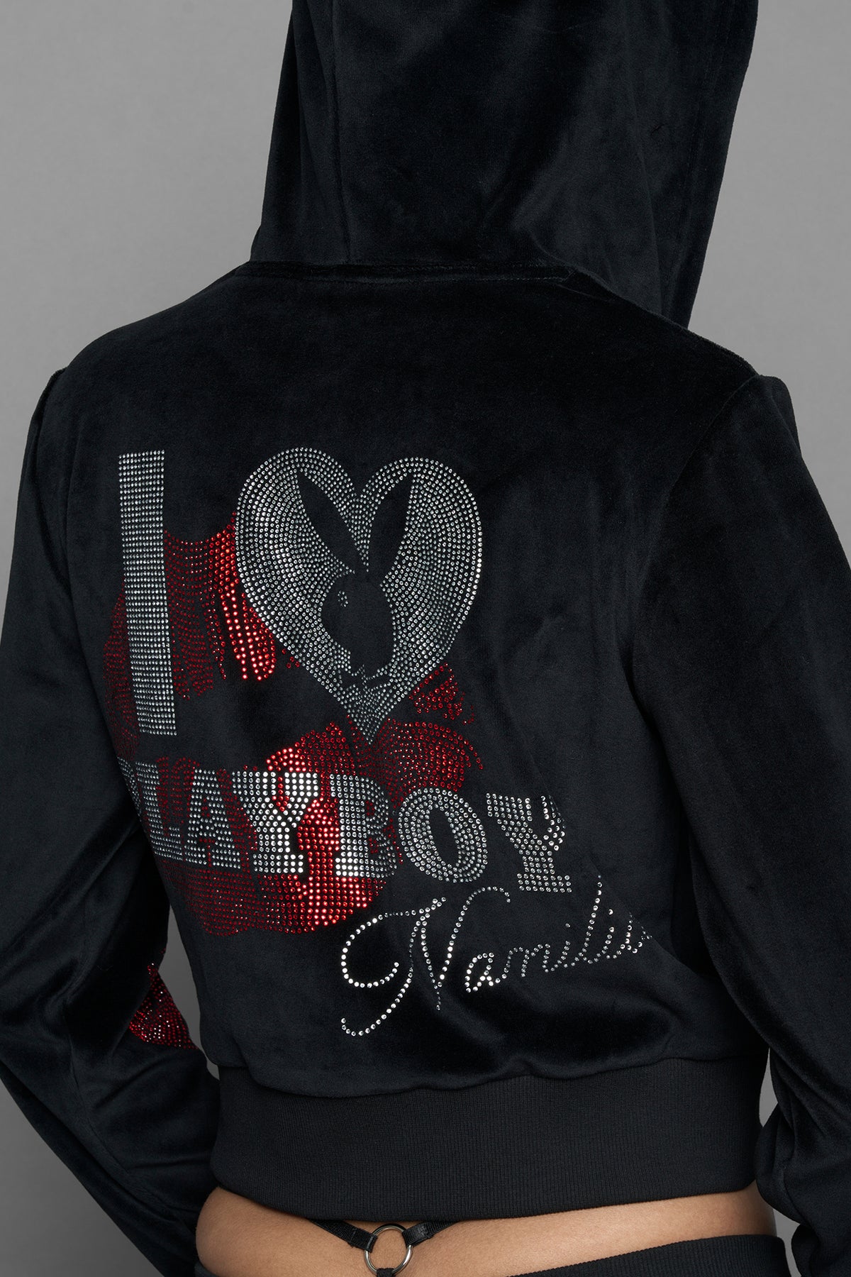 A female model wearing a black velvet zip-up hoodie with red sequined lip-print patches on the chest and sleeve, featuring white 'Namilia' embroidery on the left chest, photographed against a solid gray studio background.