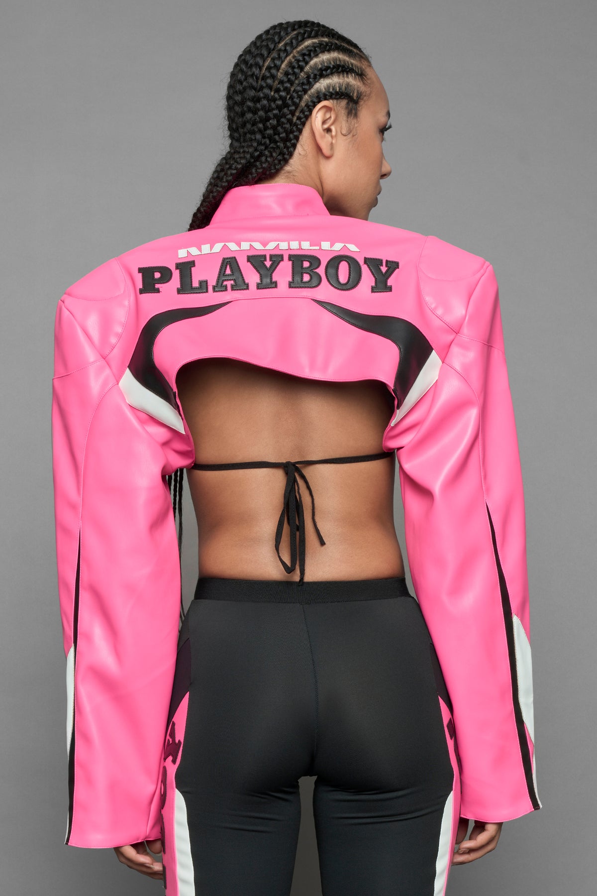 model wearing a vegan leather cropped jacket with broad shoulders in pink with black and white paneling. The jacket has PU leather embroidery of 'Namilia' and 'Playboy' with a key formed zipper pull and a rubbertag on the left breast.