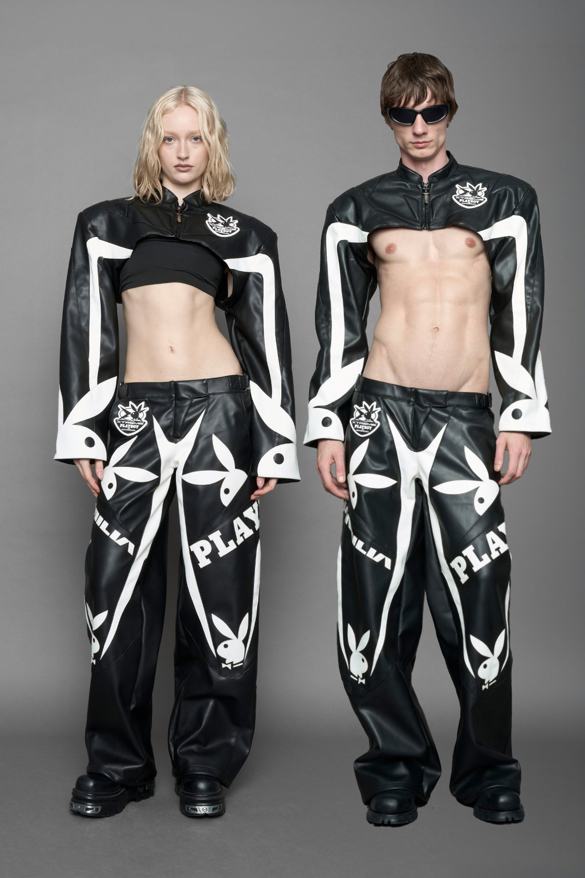 model wearing a vegan leather cropped jacket with broad shoulders in black with white paneling. The jacket has PU leather embroidery of 'Namilia' and 'Playboy' with a key formed zipper pull and a rubbertag on the left breast.