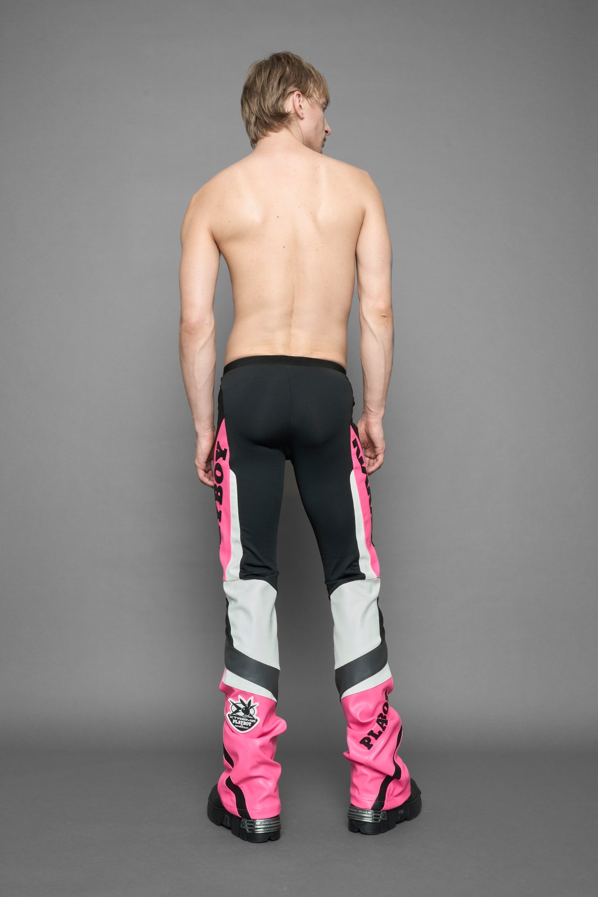 A female model wearing a pink and black form fitted Pants with PU Leather Embroidery of 'Namilia', 'Playboy'. The pants have black Mesh fabric and dark grey PU leather  panels and are slightly flared from knee to hem.