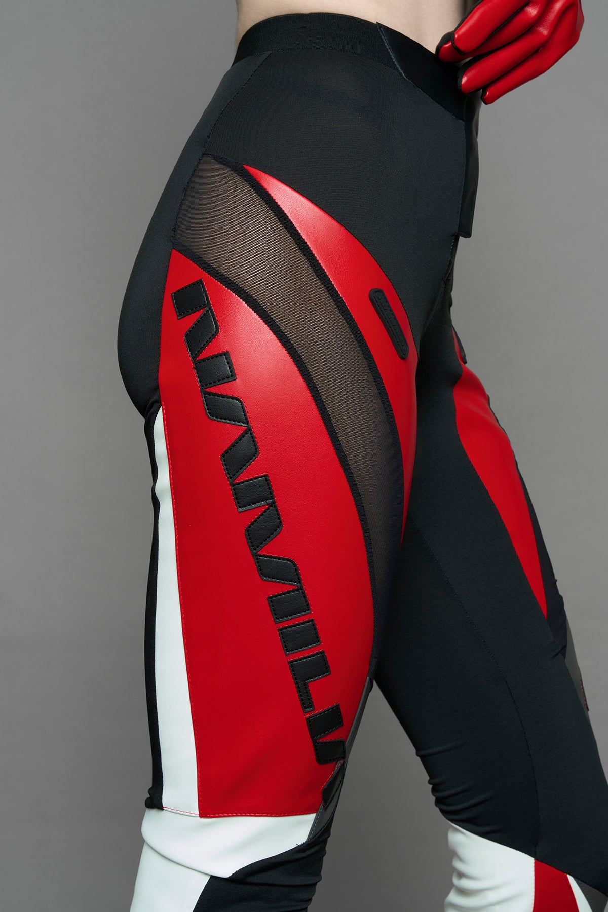 model wearing a red and black form fitted Pants with PU Leather Embroidery of 'Namilia', 'Playboy'. The pants have black Mesh fabric and dark grey PU leather  panels and are slightly flared from knee to hem