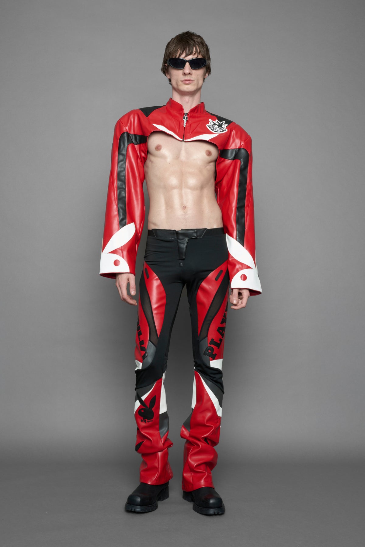 model wearing a red and black form fitted Pants with PU Leather Embroidery of 'Namilia', 'Playboy'. The pants have black Mesh fabric and dark grey PU leather  panels and are slightly flared from knee to hem