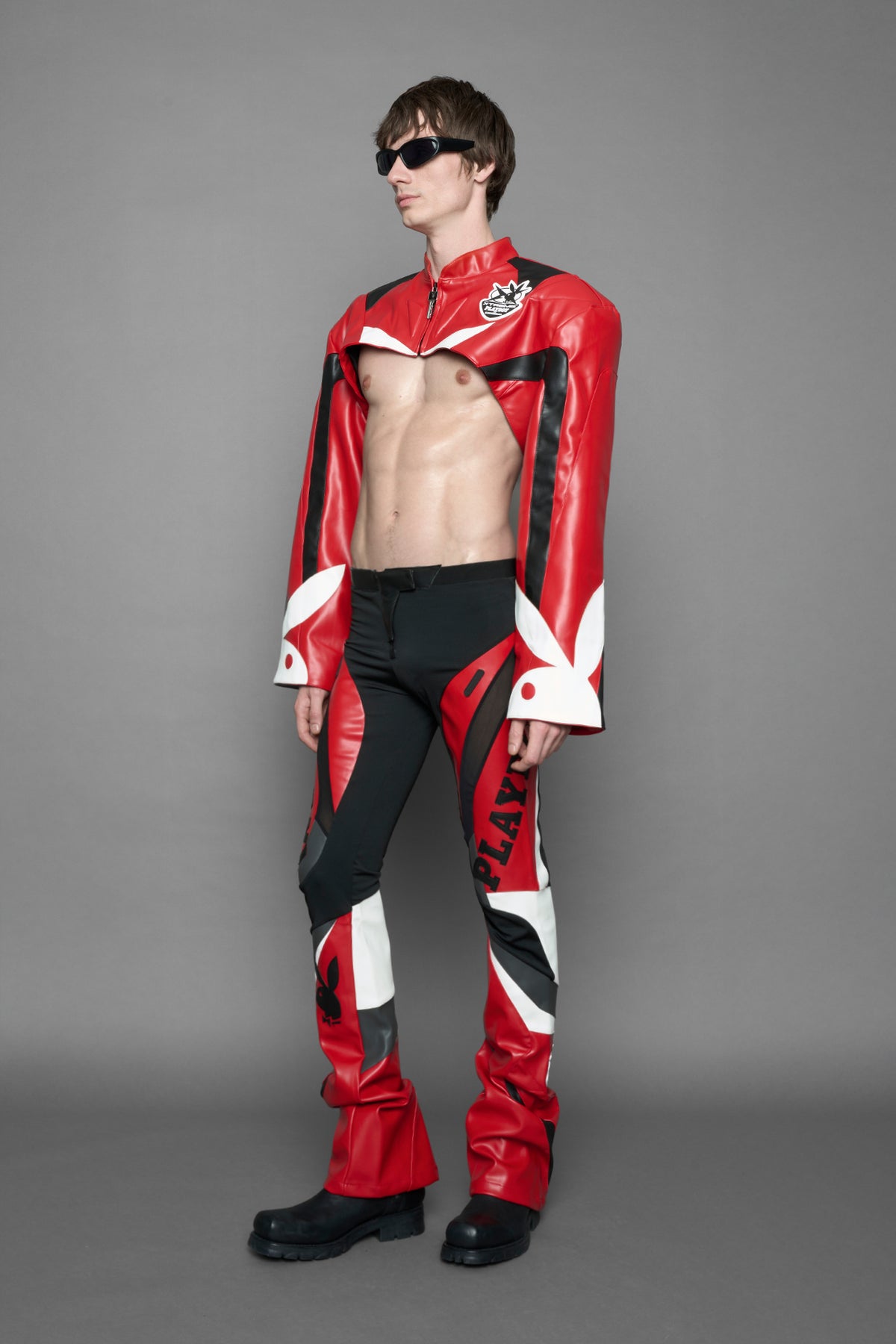 model wearing a red and black form fitted Pants with PU Leather Embroidery of 'Namilia', 'Playboy'. The pants have black Mesh fabric and dark grey PU leather  panels and are slightly flared from knee to hem