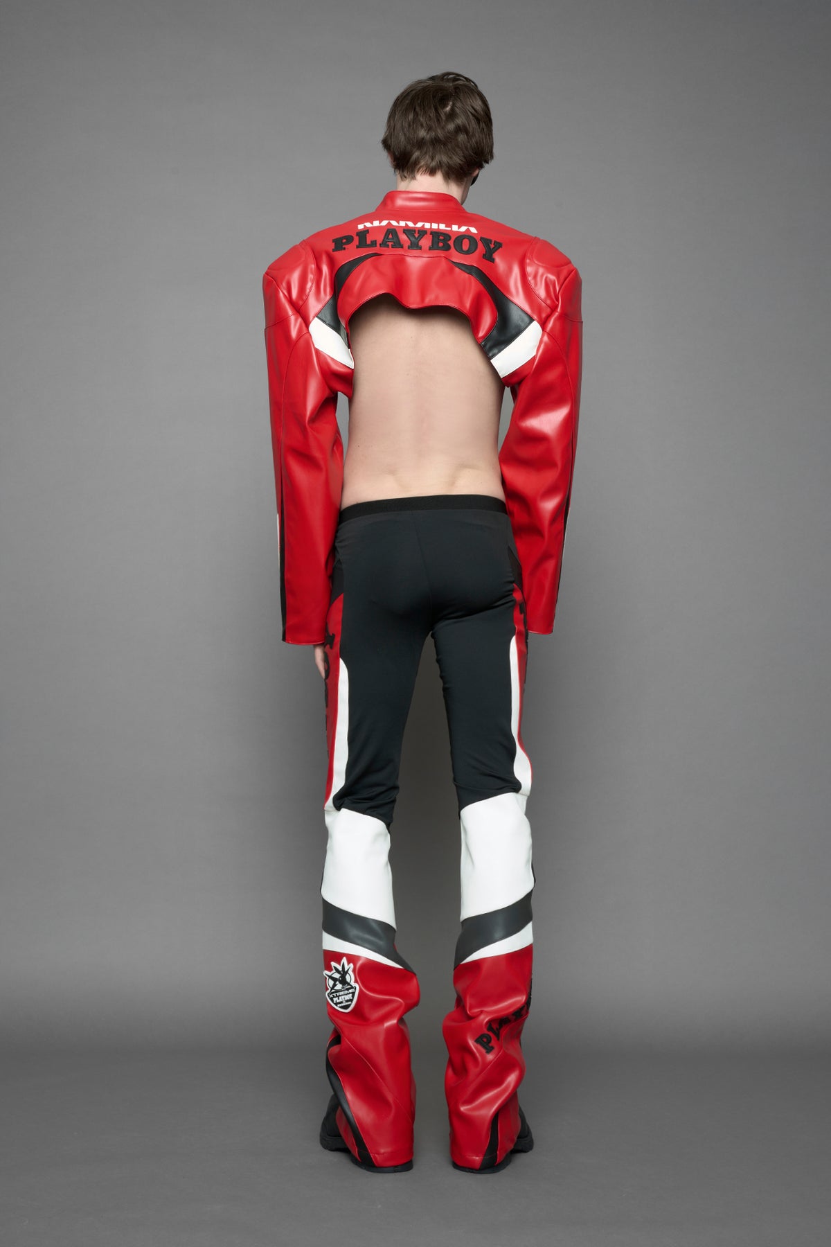 model wearing a red and black form fitted Pants with PU Leather Embroidery of 'Namilia', 'Playboy'. The pants have black Mesh fabric and dark grey PU leather  panels and are slightly flared from knee to hem