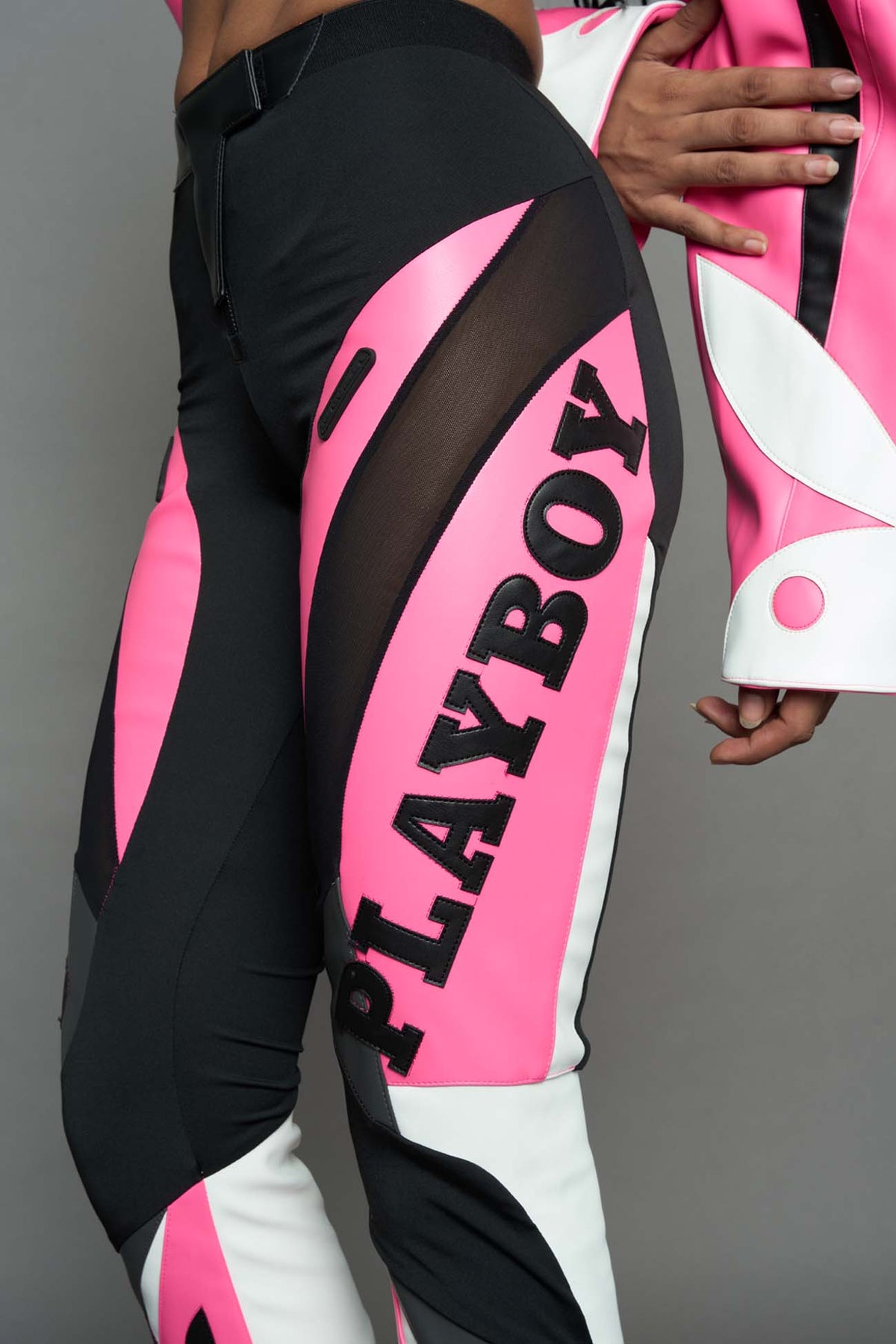 A female model wearing a pink and black form fitted Pants with PU Leather Embroidery of 'Namilia', 'Playboy'. The pants have black Mesh fabric and dark grey PU leather  panels and are slightly flared from knee to hem.