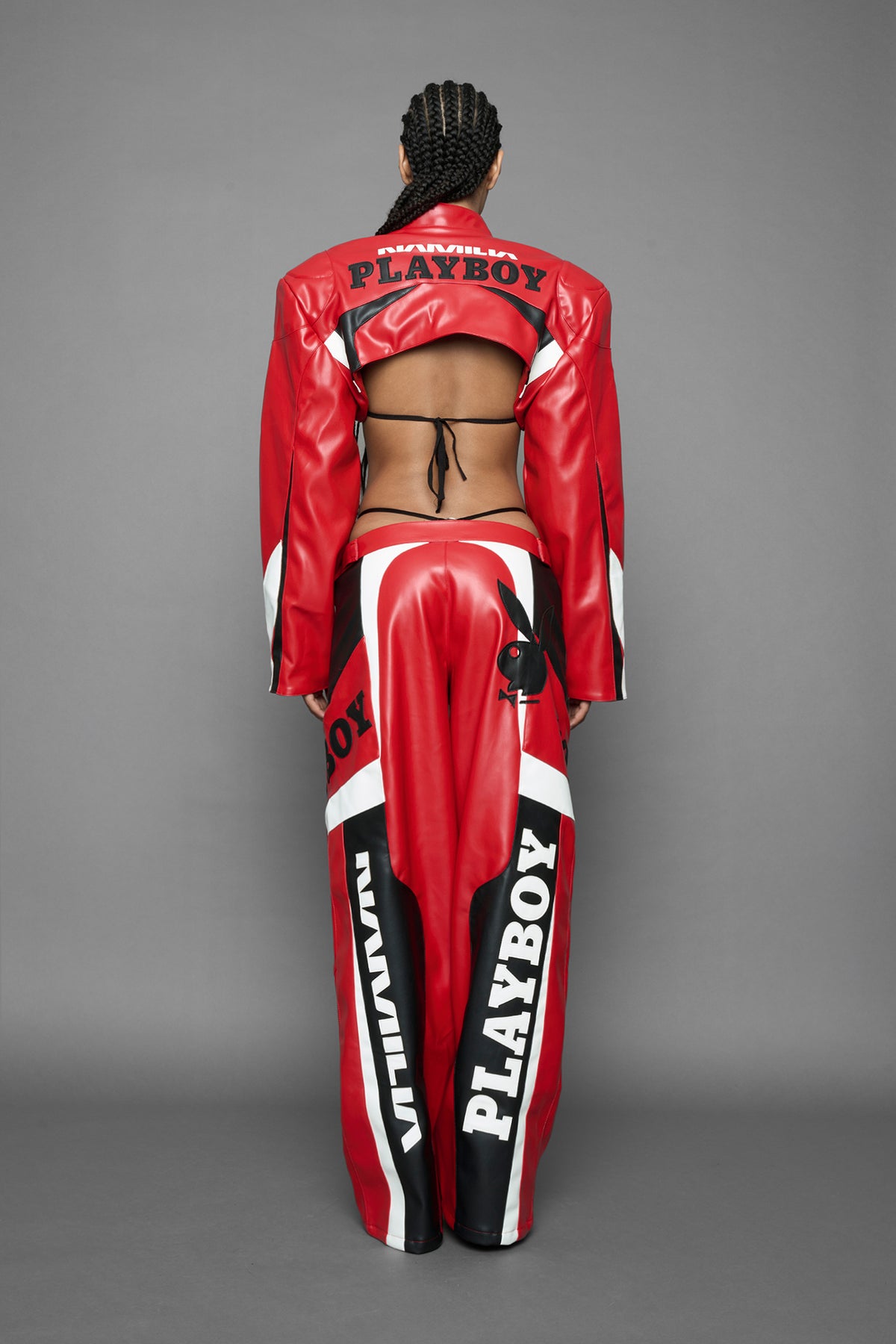 model wearing a vegan leather cropped jacket with broad shoulders in red with black and white paneling. The jacket has PU leather embroidery of 'Namilia' and 'Playboy' with a key formed zipper pull and a rubbertag on the left breast.