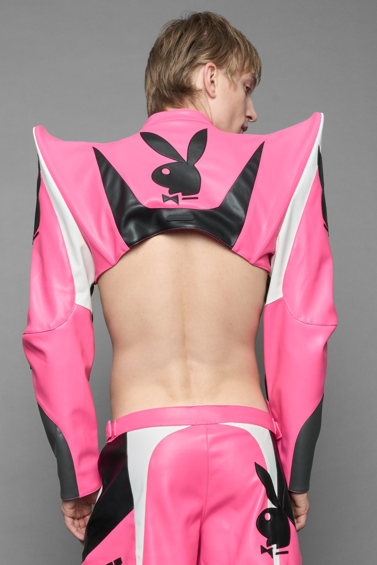  model wearing a vegan leather cropped jacket with spike silhouette shoulders in pink with black and white paneling. The jacket has PU leather embroidery of 'Namilia' and 'Playboy' with a key formed zipper pull and a rubbertag on the left breast.

