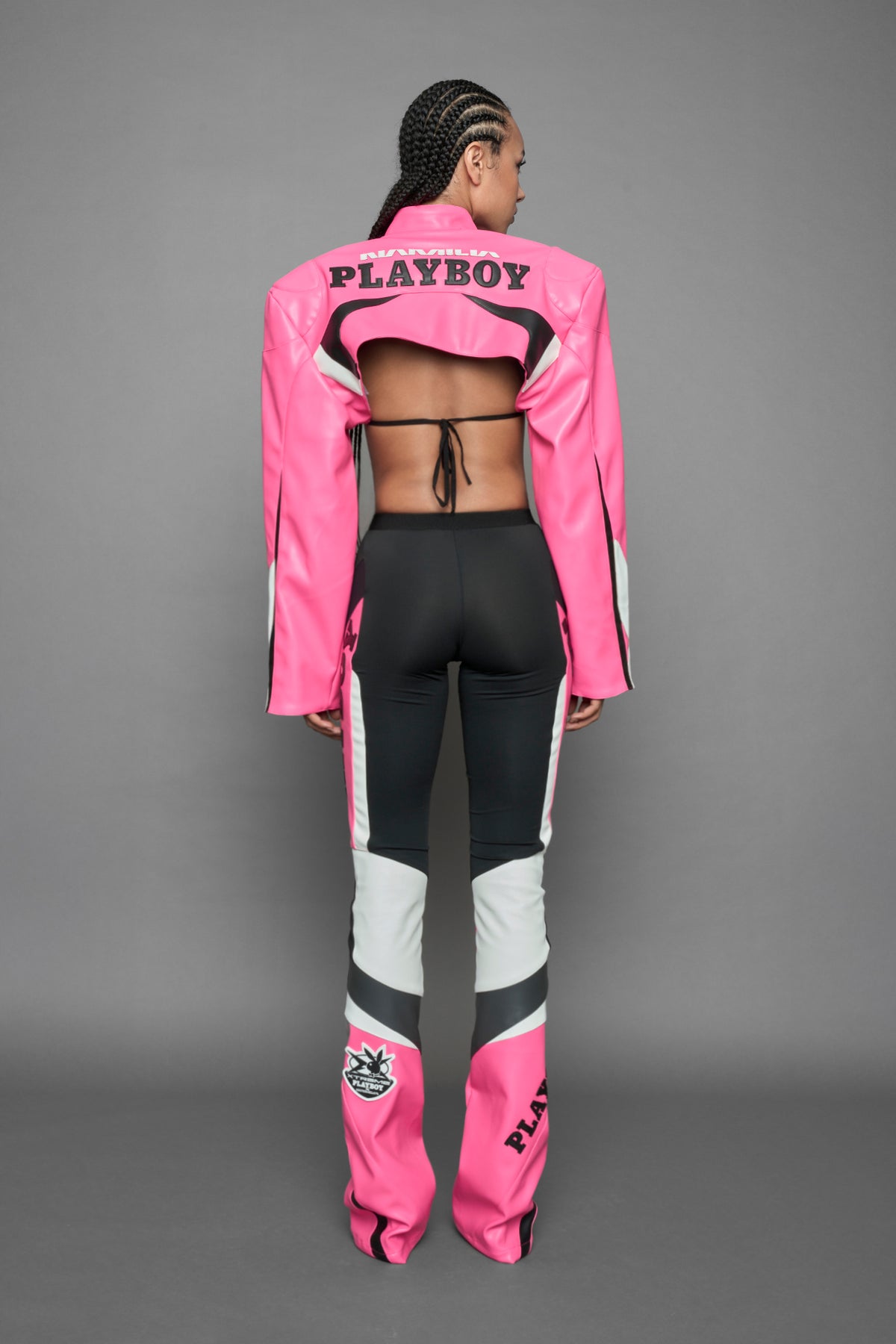 A female model wearing a pink and black form fitted Pants with PU Leather Embroidery of 'Namilia', 'Playboy'. The pants have black Mesh fabric and dark grey PU leather  panels and are slightly flared from knee to hem.