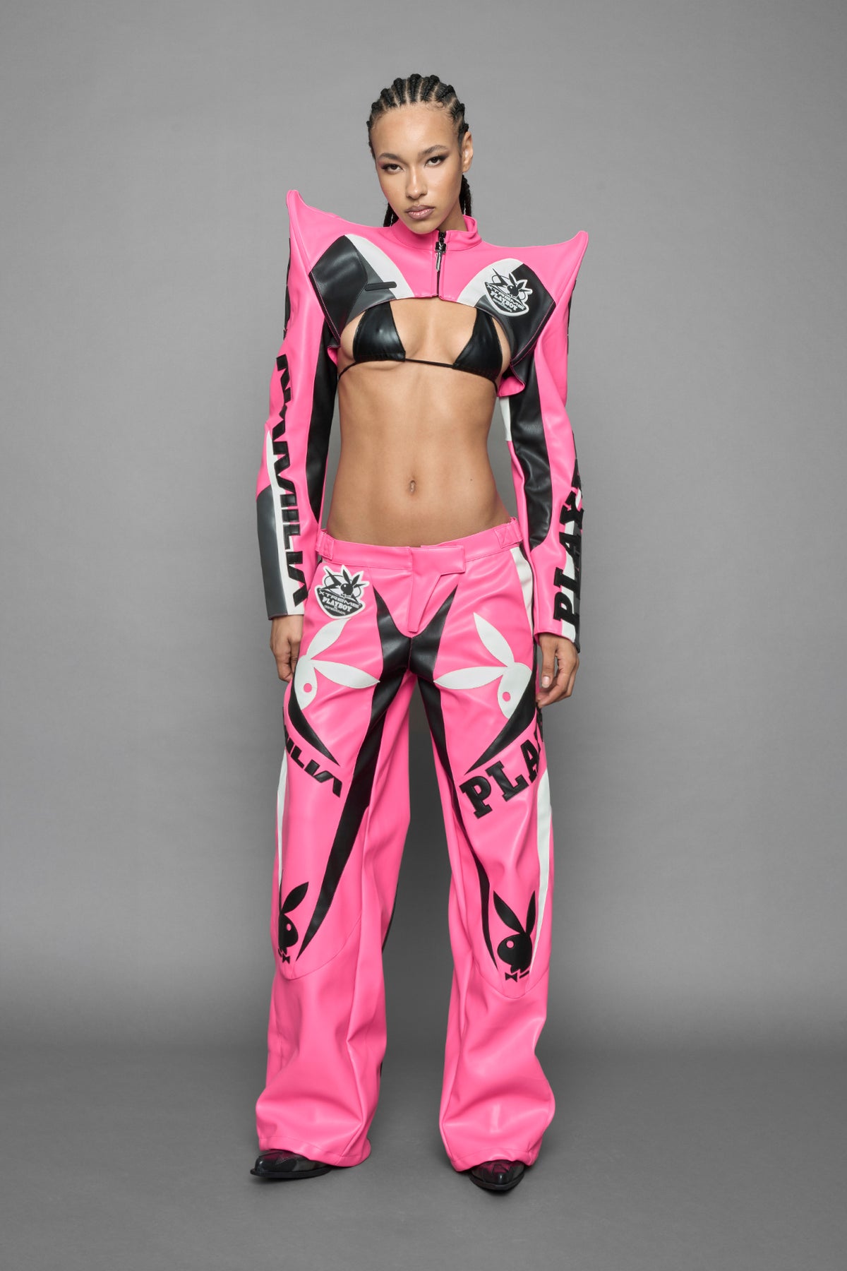  model wearing a vegan leather cropped jacket with spike silhouette shoulders in pink with black and white paneling. The jacket has PU leather embroidery of 'Namilia' and 'Playboy' with a key formed zipper pull and a rubbertag on the left breast.
