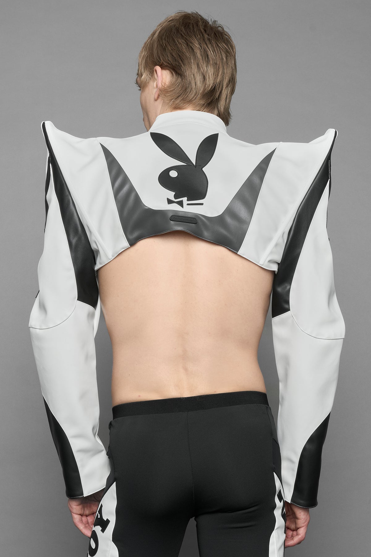 A model wearing a vegan leather cropped jacket with spike silhouette shoulders in white with black paneling. The jacket has PU leather embroidery of 'Namilia' and 'Playboy' with a key formed zipper pull and a rubbertag on the left breast.