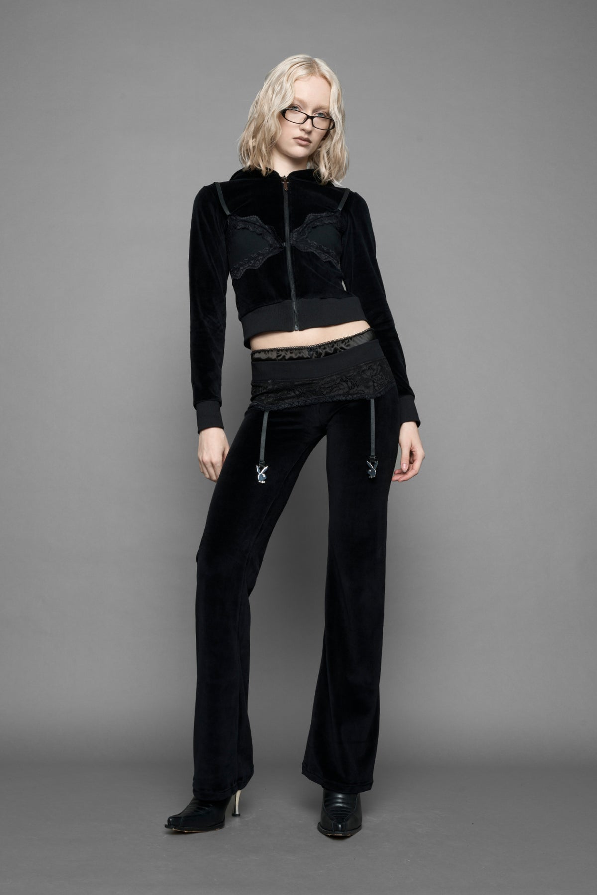 A female model wearing black velvet tracksuit pants with a lingerie black lace garter and suspenders with metal silver Bunnys 