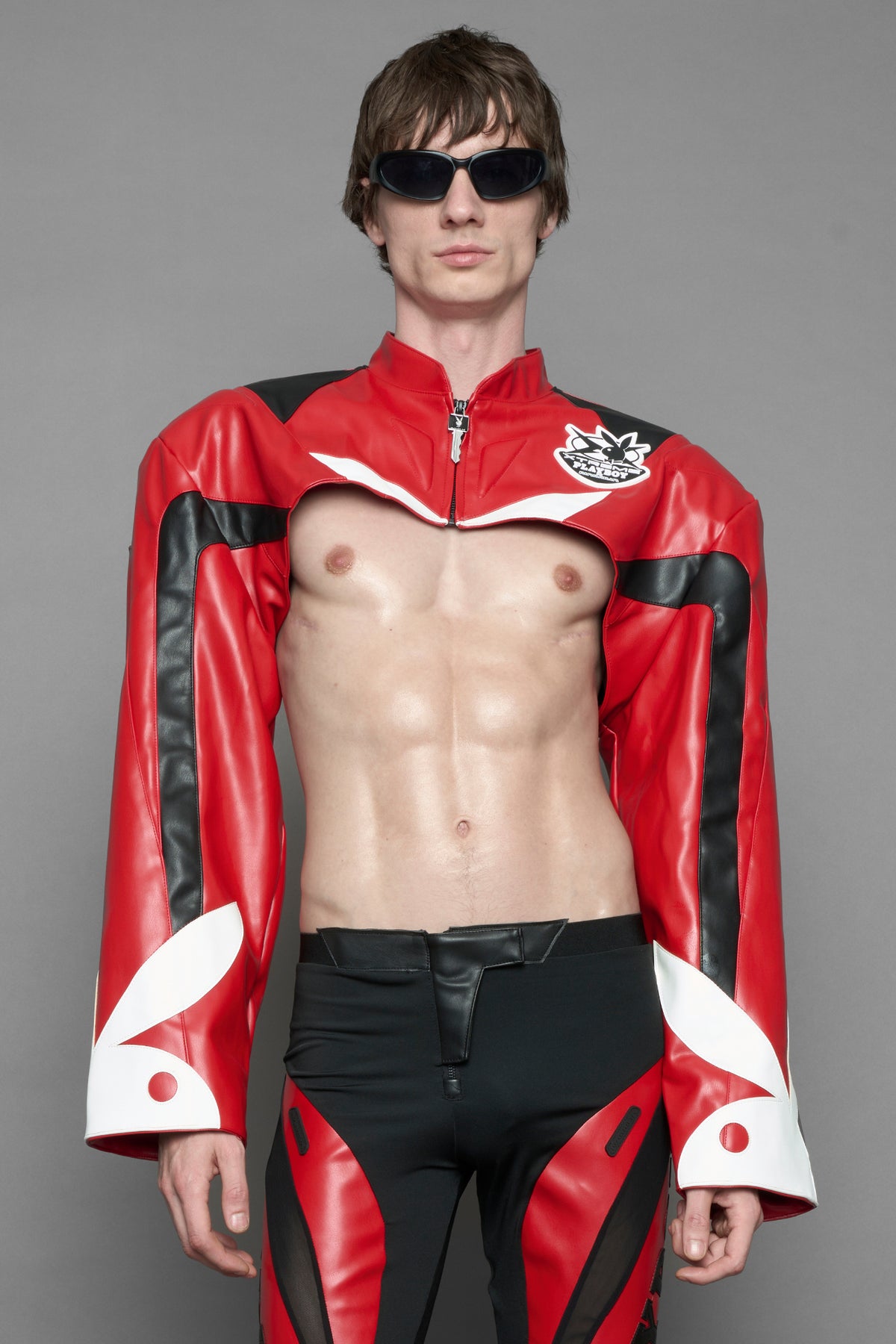 A male model wearing a vegan leather cropped jacket with broad shoulders in red with black and white paneling. The jacket has PU leather embroidery of 'Namilia' and 'Playboy' with a key formed zipper pull and a rubbertag on the left breast.