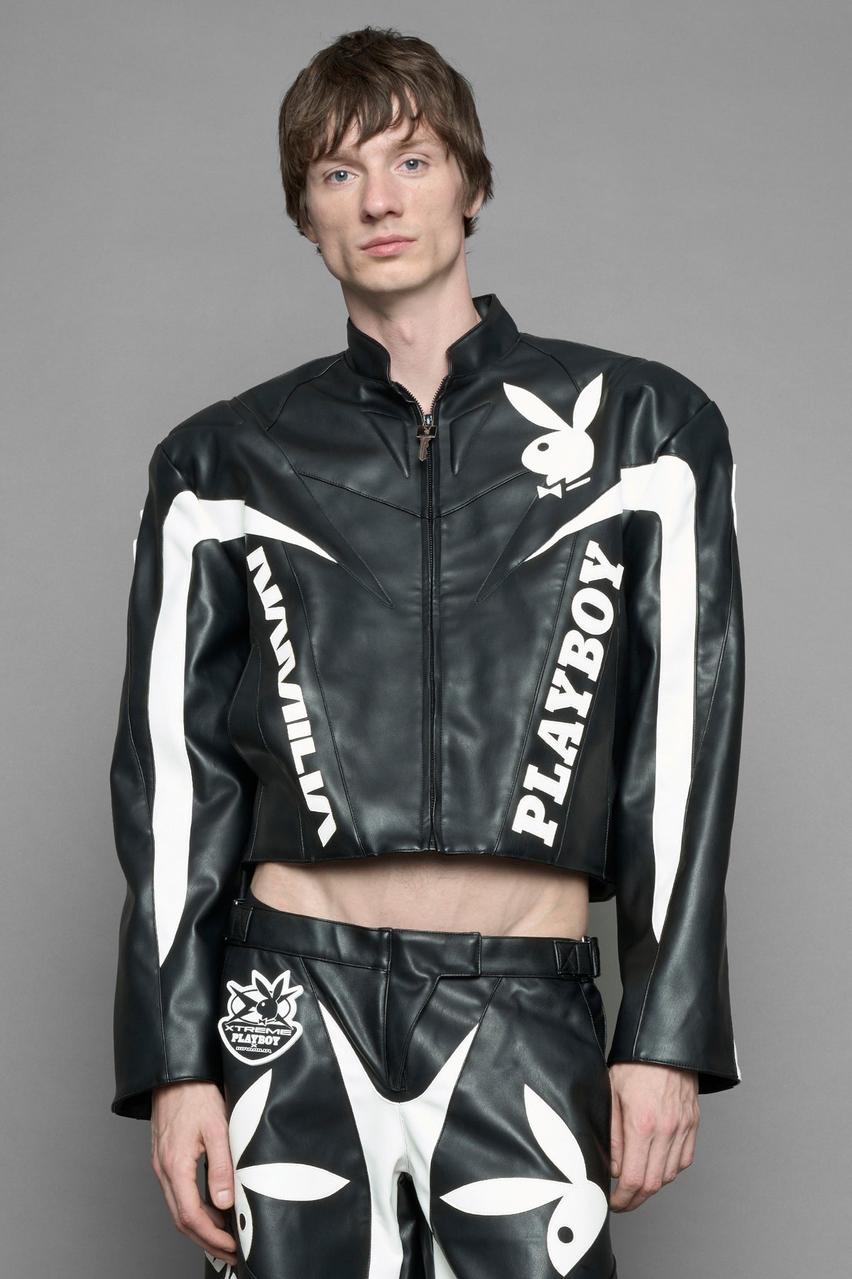 A male model wearing black vegan leather zip-up jacket with white PU leather 'Namilia' and 'Playboy' embroidery and black and white paneling. On the left chest there is a white Playboy Bunny Embroidery.
